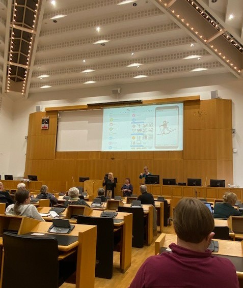 Valtuustosalissa vietettiin testiryhmäpäivää 11.5.2022. Edessä on Marjukka Nisula puhumassa ja tulkit ovat hänen vieressään. Screenillä näkyy kuva Kuvakom-sovelluksesta.