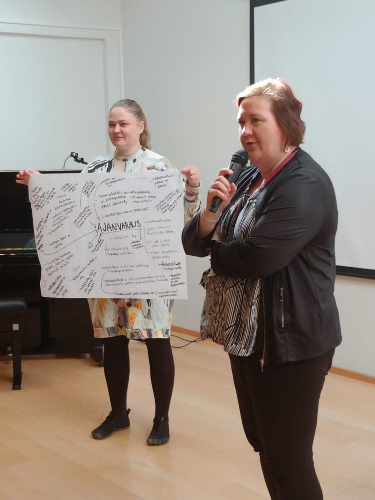 Minna Kaija-Kortelainen ja Tanja Gavrilov esittelevät ajanvaraukseen liittyviä huomioita. Minna puhuu mikrofoniin ja Tanja näyttää suurelle paperille koottuja huomioita ajanvarauksesta.