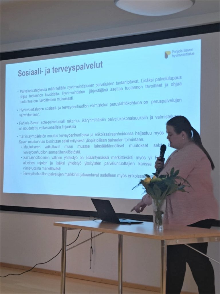 Siilinjärven kunnan sosiaali- ja terveysjohtaja Kati Kantanen kertomassa Pohjois-Savon hyvinvointialueesta. Hän puhuu mikrofoniin ja näyttää esittelydioja.