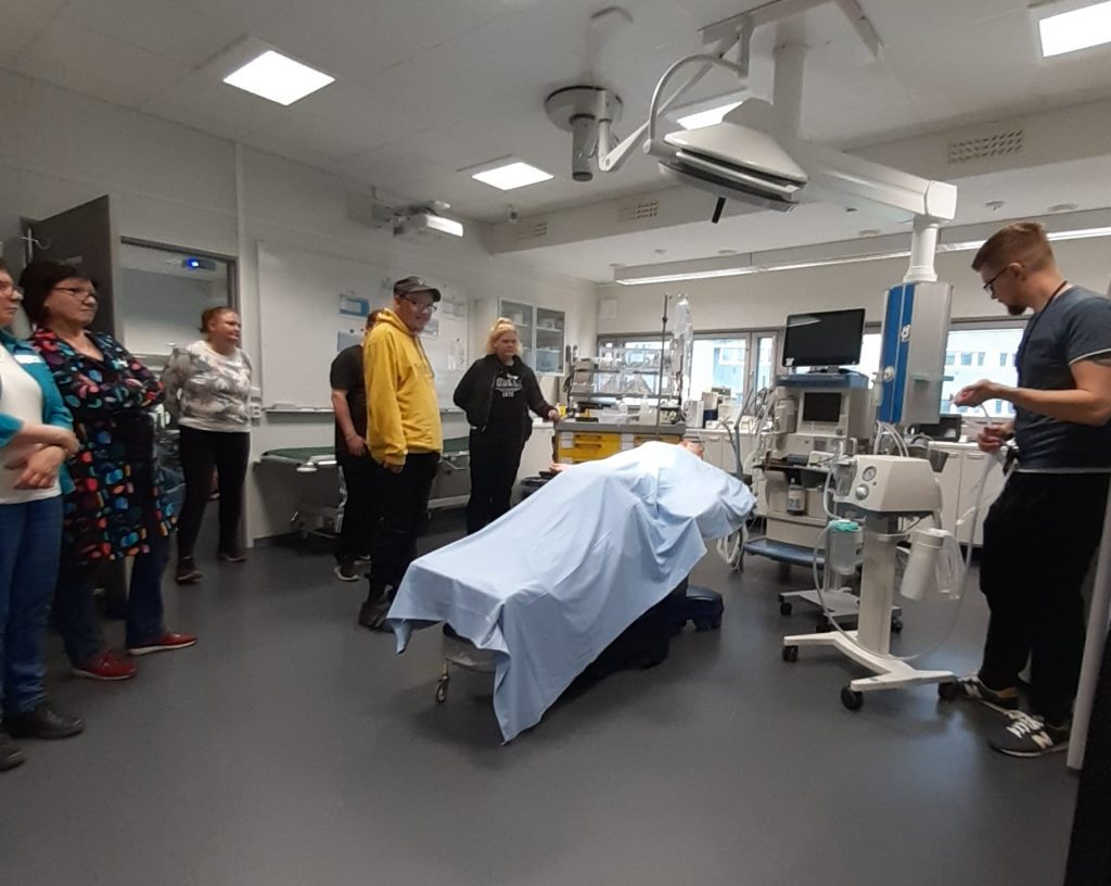 Ryhmä on kokoontunut simulaatiotilassa kuuntelemaan asiantuntijan selkokielistä esitystä sairaalan hoitovälineistä.